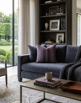 Dark gray sofa with a purple and striped purple Mayan pillow cover centered, accented by a dark gray knit throw and a light wood coffee table with books and a ceramic mug.