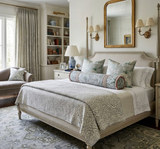 Traditional bedroom with a beige upholstered bed featuring sage floral pillows and a blue-green bolster, flanked by matching sconces and a gold-framed mirror above.