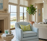 Light blue armchair with a pistachio green Mille Feuilles pillow cover featuring a leafy pattern, set near large windows with ocean views and beige furnishings.