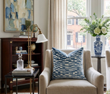 Beige armchair with a large marine blue and white patterned velvet pillow in a bright room with a wooden cabinet, gold-toned lamp, and a blue and white floral vase on a side table.