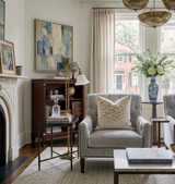 Light gray tufted armchair with a tan brown velvet pillow featuring a geometric pattern, set in a softly lit living room with neutral tones and large windows.