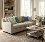 Neutral beige sofa with five decorative pillows including blue, green, and patterned designs, placed in a bright living room with wooden floor and a coffee table with books and a vase.