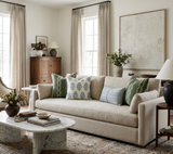 Light beige sofa with assorted patterned throw pillows in muted blue, green, and gray, set in a bright living room with a marble coffee table and neutral decor.