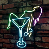 Neon sign depicting a reclining figure in green and yellow neon lines inside a bright blue martini glass against a dark red brick wall background.