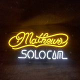 Neon sign with yellow cursive text 'Mathews' above white block letters spelling 'SOLOCOM' on a dark background.