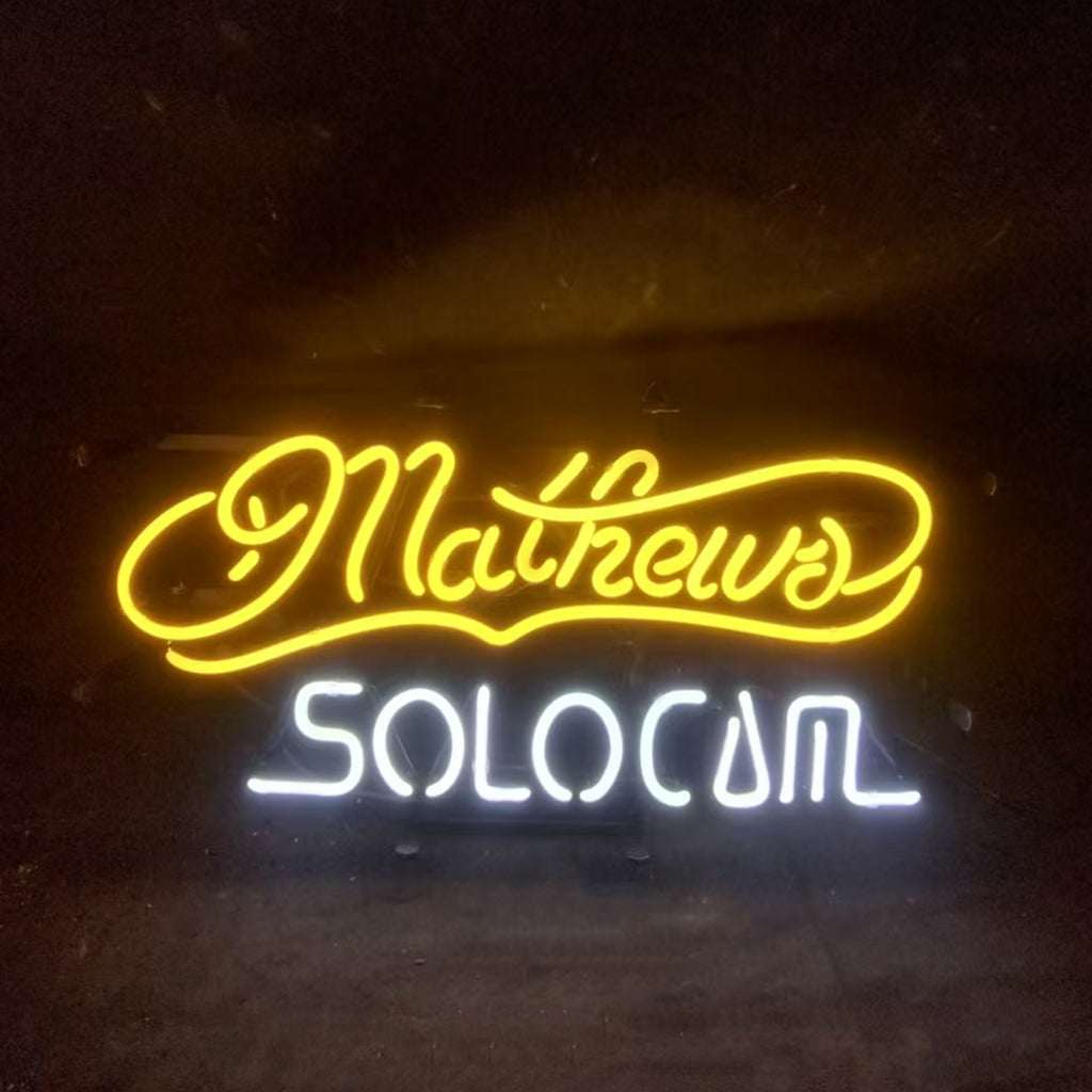 Neon sign with yellow cursive text 'Mathews' above white block letters spelling 'SOLOCOM' on a dark background.