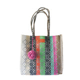Colorful woven tote bag with vertical stripes in gray, orange, green, pink, and brown, featuring diamond patterns and braided handles with a pink pom-pom accent.