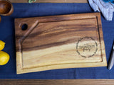 Medium rectangular live edge wood cutting board with a juice groove, rounded corner hole for hanging, and personalized engraving reading 'the Thompson family est. 2021'