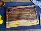 Rectangular live edge wood cutting board with hanging hole and juice groove, engraved with "John & Olivia 9.20.2020" on a blue textile background.