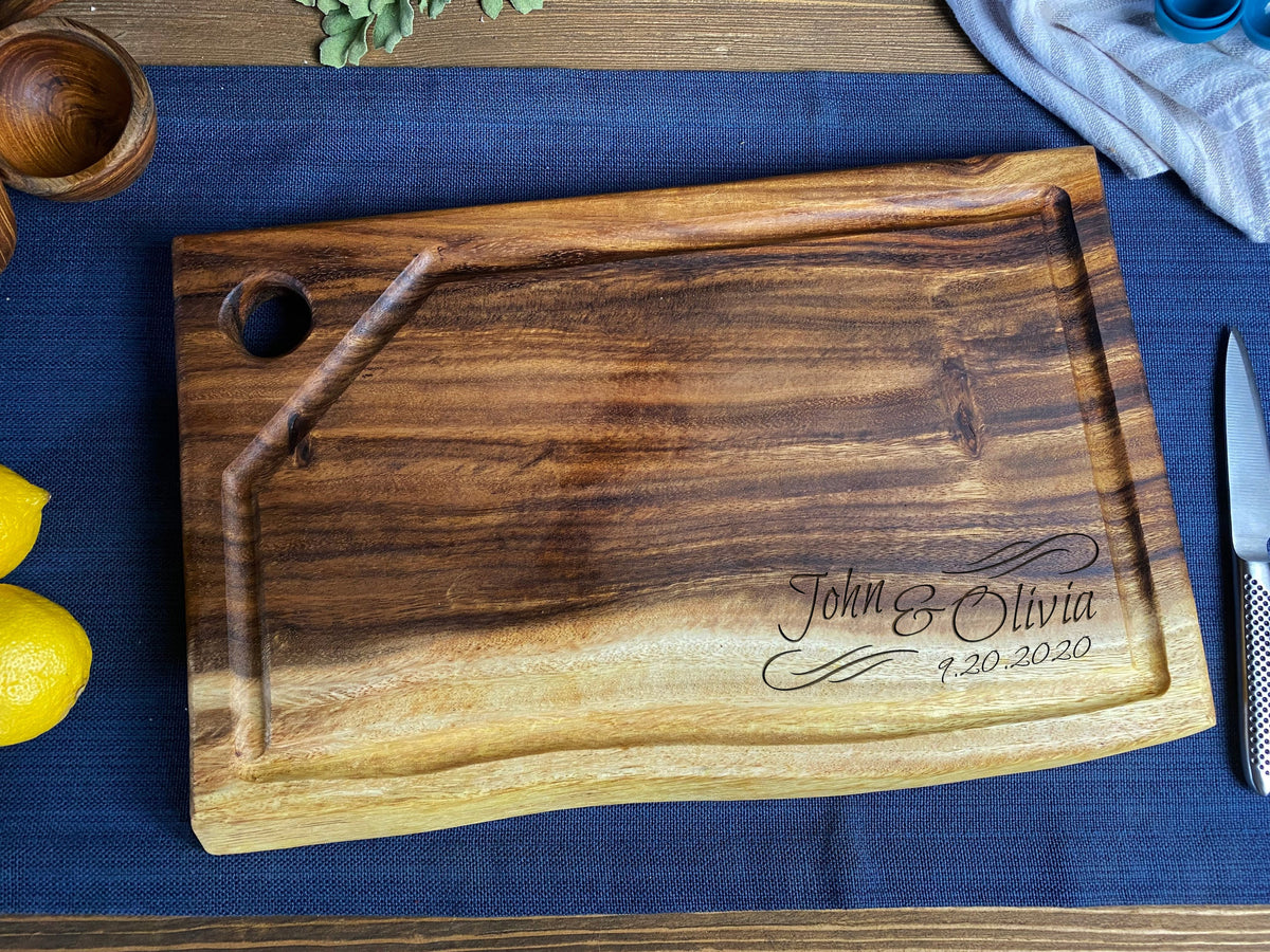 Rectangular live edge wood cutting board with hanging hole and juice groove, engraved with "John & Olivia 9.20.2020" on a blue textile background.