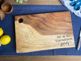 Rectangular wood cutting board with live edges, a round hanging hole, and engraved "EST. the 2011 THOMPSON family" text, placed on blue fabric on wooden table.
