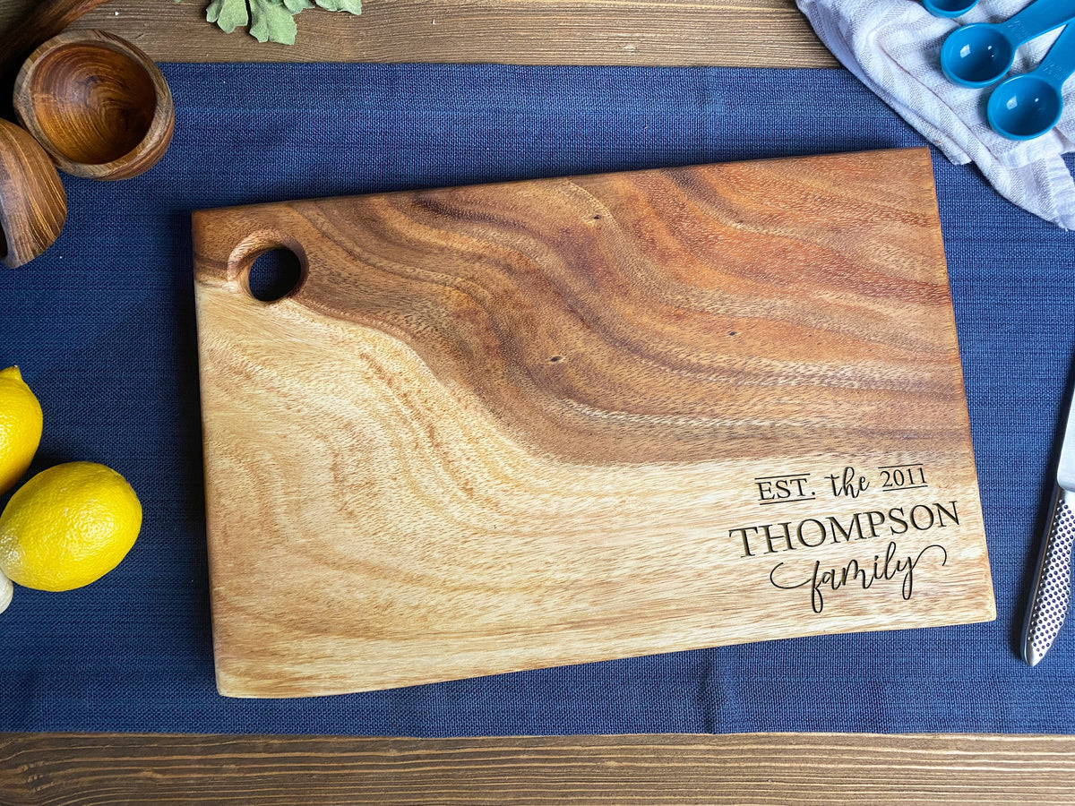 Rectangular wood cutting board with live edges, a round hanging hole, and engraved "EST. the 2011 THOMPSON family" text, placed on blue fabric on wooden table.