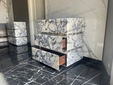 Freestanding vanity unit with three marble drawers featuring white stone and dark gray veining, shown with one drawer partially open against a matching marble wall and floor.