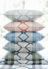 Stack of six rectangular pillows with geometric diamond patterns in alternating colors including blue, rust, dark gray, and olive green, set against a bright window background.