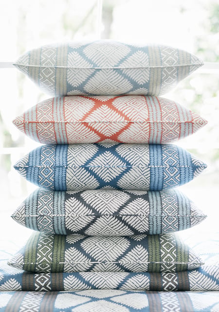 Stack of six rectangular pillows with geometric diamond patterns in alternating colors including blue, rust, dark gray, and olive green, set against a bright window background.