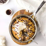Mini stainless steel frying pan with a baked dessert topped with whipped cream and chocolate chips, resting on a round wooden trivet with a white cloth nearby.