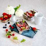 Two small silver mini roasters filled with fruit-topped desserts, surrounded by fresh strawberries, raspberries, blueberries, mint leaves, a white coffee cup, and a small white vase with white flowers on a light surface.