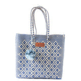 Woven tote bag with blue and natural beige diamond pattern, featuring twisted handles and a blue pom-pom accessory attached near the top.