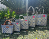 Five woven bags in blue and cream diamond patterns with brown or black handles, labeled Small through Super Tote, displayed outdoors on grass with greenery behind.