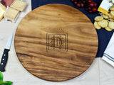 Round wooden charcuterie board with a dark grain pattern and a decorative letter D engraved in the center, placed on a light wood table surrounded by snacks.