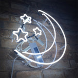 White neon light sign shaped like a crescent moon with three stars, mounted on a clear circular background against a brick wall.