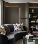 Curved dark gray velvet sofa with assorted patterned and solid pillows in soft pink, beige, and multicolor, positioned near wooden paneling with integrated lighting and a glass coffee table.