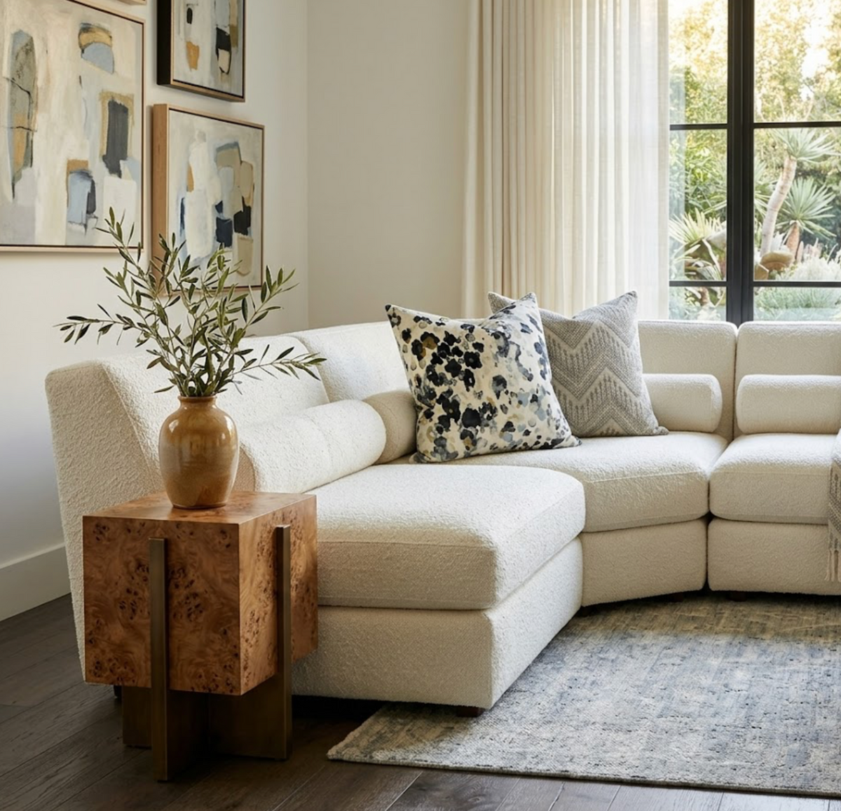 Curved white textured sectional sofa with patterned pillows, next to a cube-shaped wooden side table holding a brown vase with leafy branches, by large window with long sheer curtains.