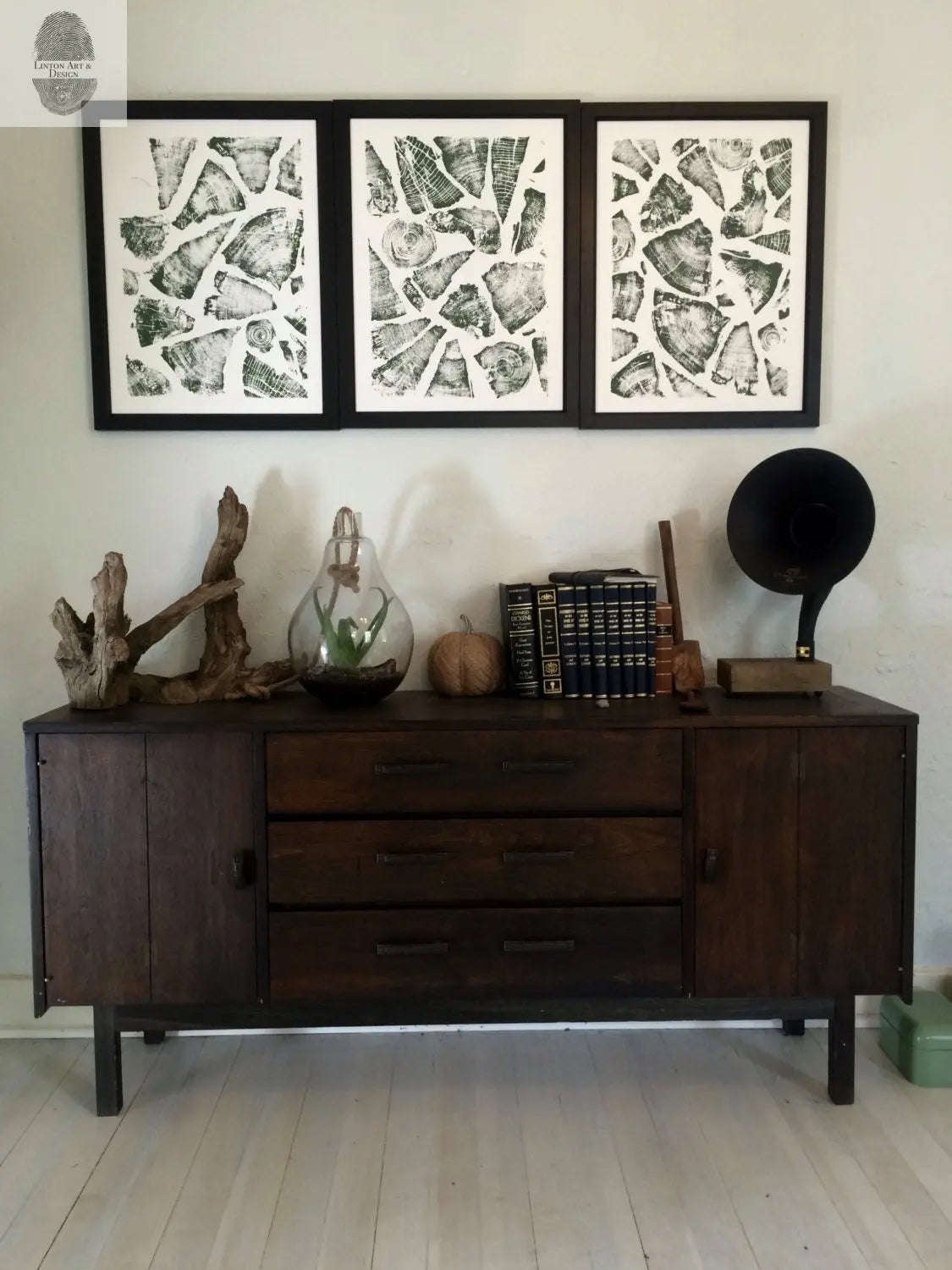 Three framed black and white natural pattern collages arranged horizontally above a dark wood sideboard with eclectic decor including driftwood, a glass terrarium, books, and a vintage gramophone.