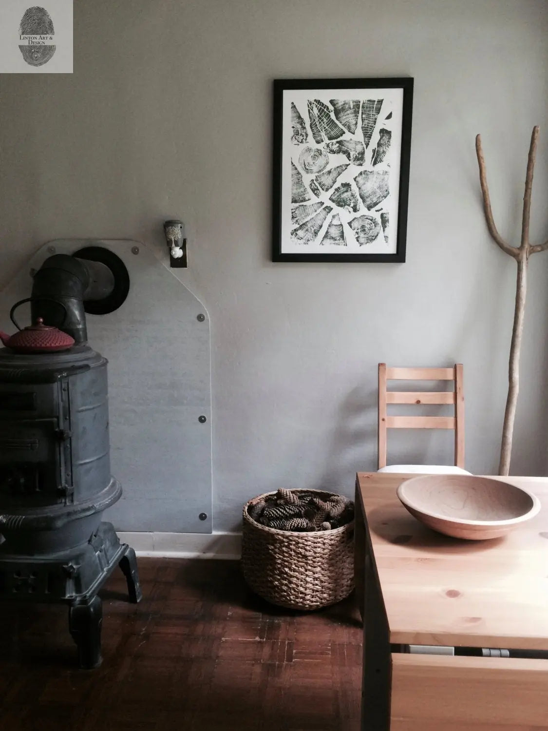 Rustic interior with black cast iron stove, wicker basket filled with pinecones, wooden table with shallow bowl, and black-framed natural pattern collage print on gray wall.