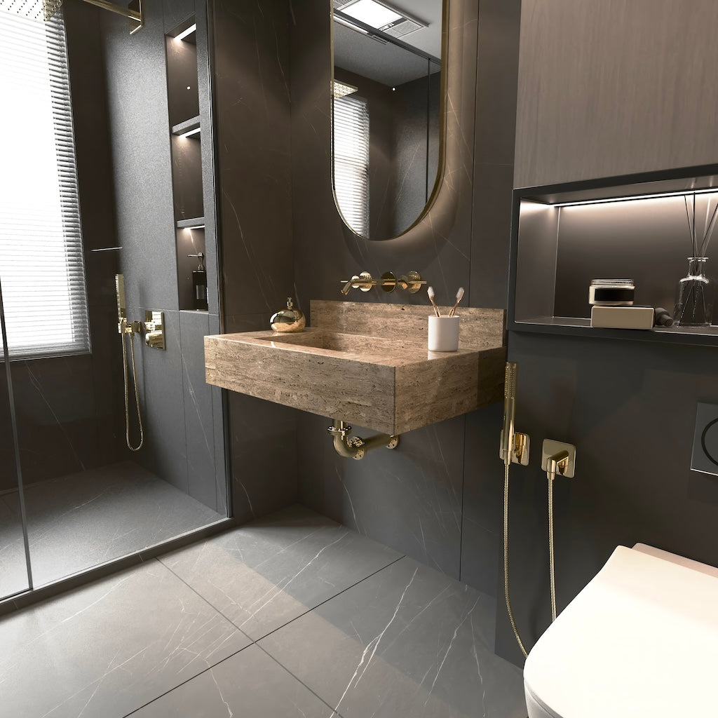 Wall-mounted rectangular travertine stone sink with integrated backsplash and brass fixtures in a modern bathroom with dark gray tiles and a large rounded mirror.