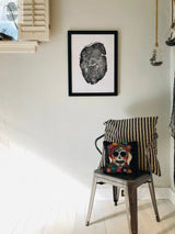 Black and white detailed print of a round wood cross-section framed on a white wall above a metal chair with a black and white patterned blanket and colorful skull pillow.