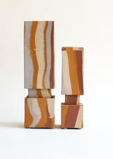 Pair of tall, rectangular ceramic vases with earthy vertical stripes in beige, rust, and mustard yellow, each resting on a square matching base against a white background.