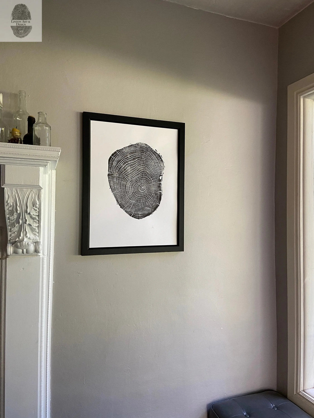 Black and white framed wood cross-section print with concentric rings mounted on a light gray wall above tufted dark blue seating near a window and white ornate mantel.