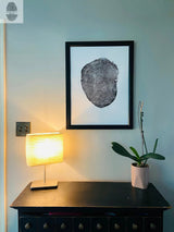 Black-and-white framed print of a tree cross-section with growth rings on a pale blue wall above a black wooden table holding a lit cream lamp and a potted orchid with green leaves.