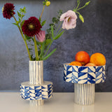 Two ceramic vessels with blue geometric and vertical line patterns; one tall vase holding flowers and one elevated bowl containing peaches, against a gray backdrop.