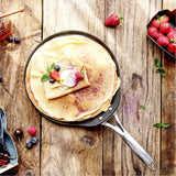 Flat crepe on a black pan with a silver fixed handle, topped with cream, fresh berries, mint, and a dusting of powdered sugar on a rustic wooden table.