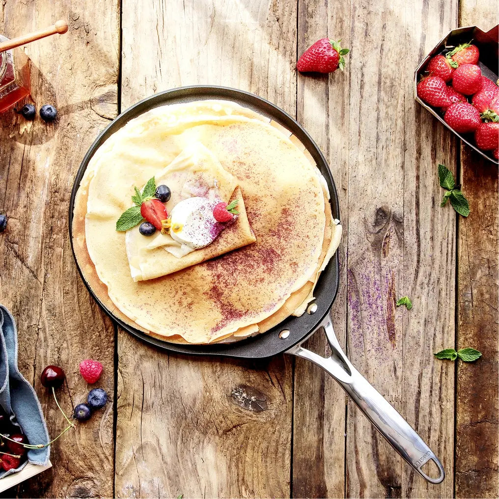 Flat crepe on a black pan with a silver fixed handle, topped with cream, fresh berries, mint, and a dusting of powdered sugar on a rustic wooden table.