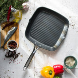 Square non-stick grill pan with ridged cooking surface and black handle, surrounded by yellow and red bell peppers, spices, olive oil, herbs, and garlic on a white textured surface.