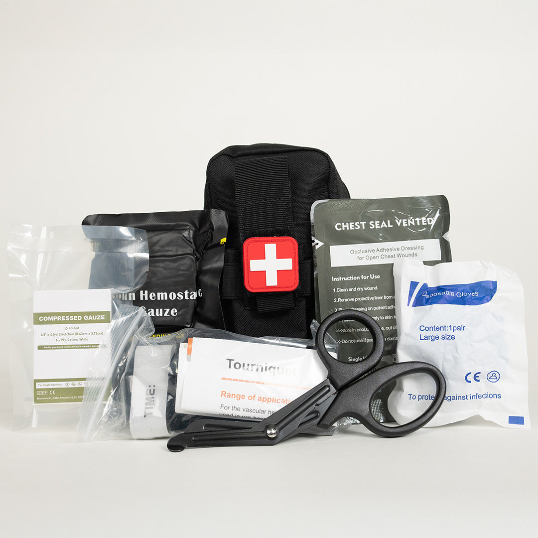 Black compact first aid pouch marked with a red cross, surrounded by medical supplies including hemostatic gauze, chest seal, tourniquet, trauma shears, and gloves on a white background.