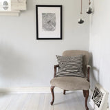 Black and white framed oak tree ring print hanging on a light gray wall above a beige upholstered wooden chair with a black and white houndstooth pillow, white wooden floor beneath.