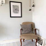 Black and white oak tree ring print in a black frame hangs on a light gray wall above a beige upholstered wooden chair with a black and white houndstooth pillow, near two hanging glass terrariums.