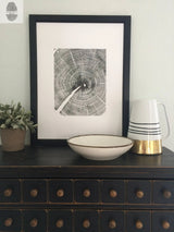 Black and white framed print of oak tree rings mounted on a light wall above a dark wooden drawer chest, accompanied by a potted plant, bowl, and striped pitcher.