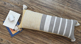 Long rectangular throw pillow with vertical stripes in beige, white, and grey, adorned with white tassels at each corner, placed on wood-textured surface with papers beneath.