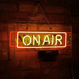 Rectangular neon sign with orange border and yellow 'ON AIR' text hanging on a brick wall with an attached power unit beneath.