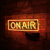 Rectangular neon sign with orange outline and yellow letters spelling 'ON AIR' mounted on a clear acrylic backing against a brick wall at night.