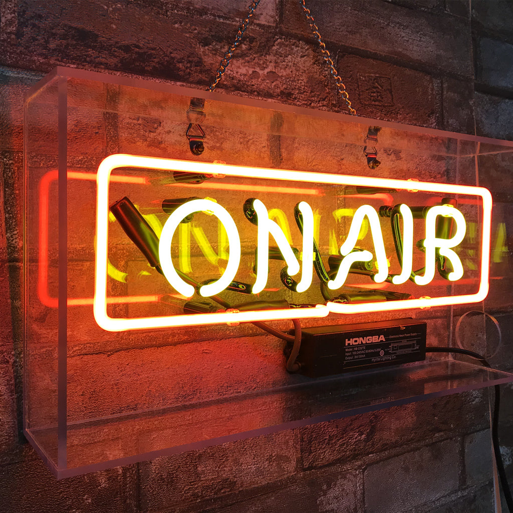 Clear acrylic box with glowing orange neon sign spelling 'ON AIR' mounted on a rustic brick wall, chain hanging from top for suspension.