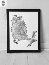 Black and white tree ring print with distinct growth rings and bark texture on white background, framed in black and leaning against a white wall on light wood floor.