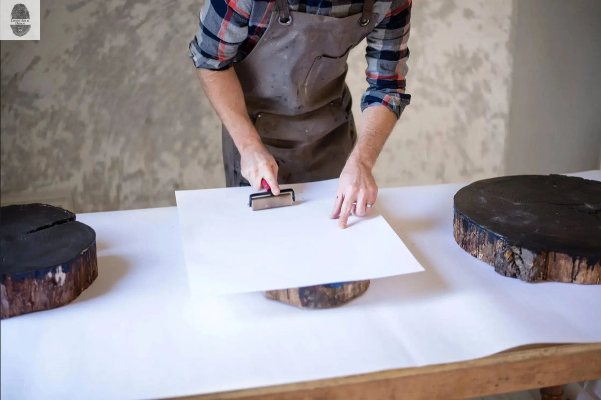 Person in plaid shirt and apron printing on white paper placed over a blackened wood slice using a brayer, with two additional dark wood slices on a white-covered workspace.