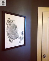 Black and white print of a detailed tree ring pattern framed in dark wood, hanging on a gray wall next to a beige door with a vintage metal doorknob.