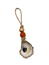 Ornament featuring a white and gold oyster shell with a black center, suspended from jute twine with a gold and a reddish-brown bead above it.